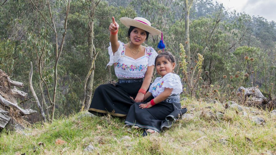 Madre señala con la mano hacia arriba mientras su hija mira con curiosidad. Están vestidas con trajes tradicionales de Otavalo, Ecuador.