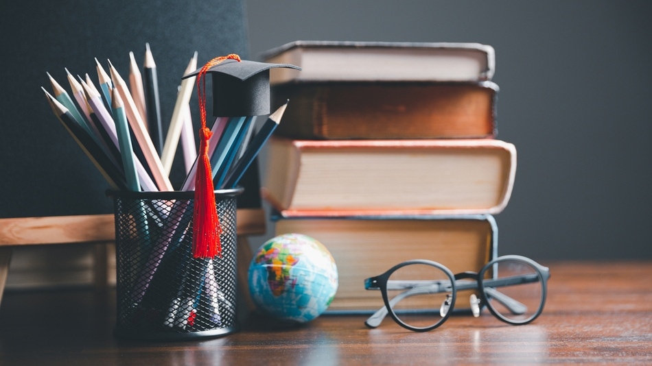 Gorra de graduación sobre lápices en un cubilete, globo terráqueo, libros y unas gafas sobre una mesa de madera